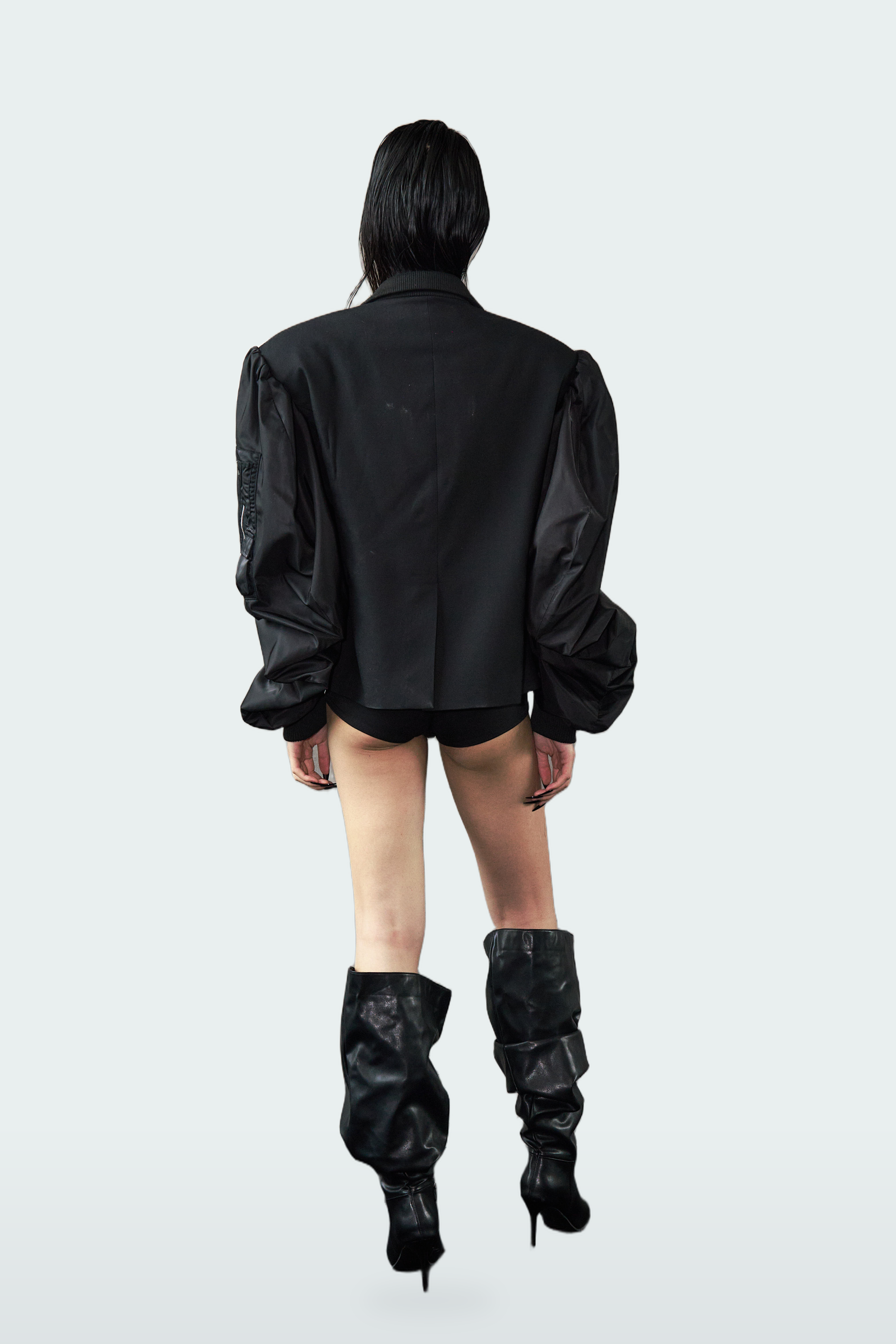 A model shown from the back wearing a black jacket with dramatic puffed balloon sleeves, black shorts, and black high-heeled boots, set against a solid light gray background.
