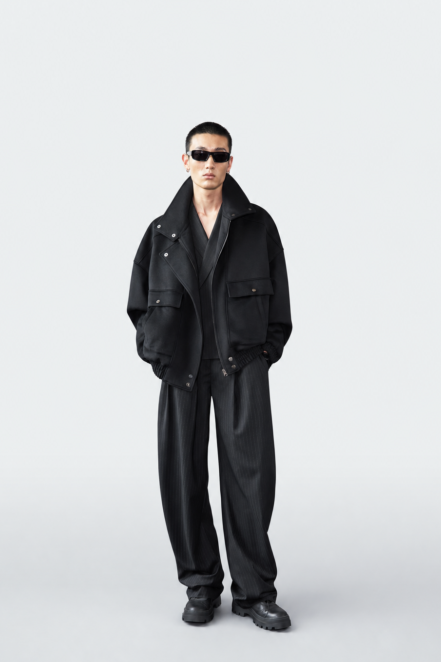 A model stands against a light gray studio background wearing a black oversized jacket, matching black trousers, black shoes, and black sunglasses.