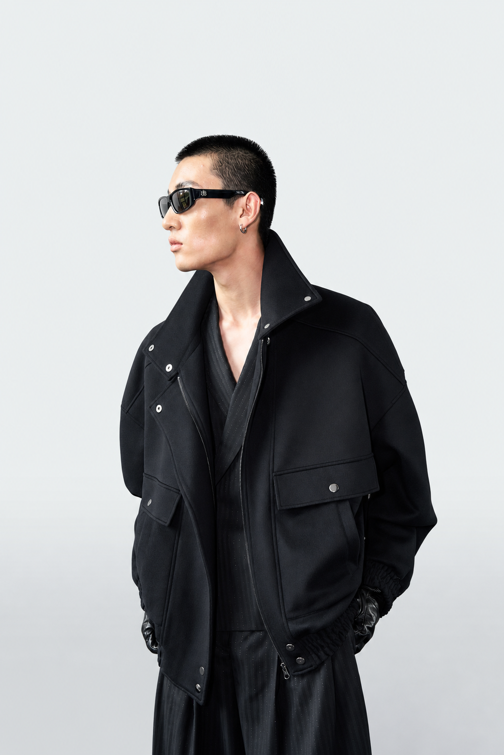 A model wearing a black oversized jacket, black sunglasses, and a black shirt, standing against a light gray studio background.