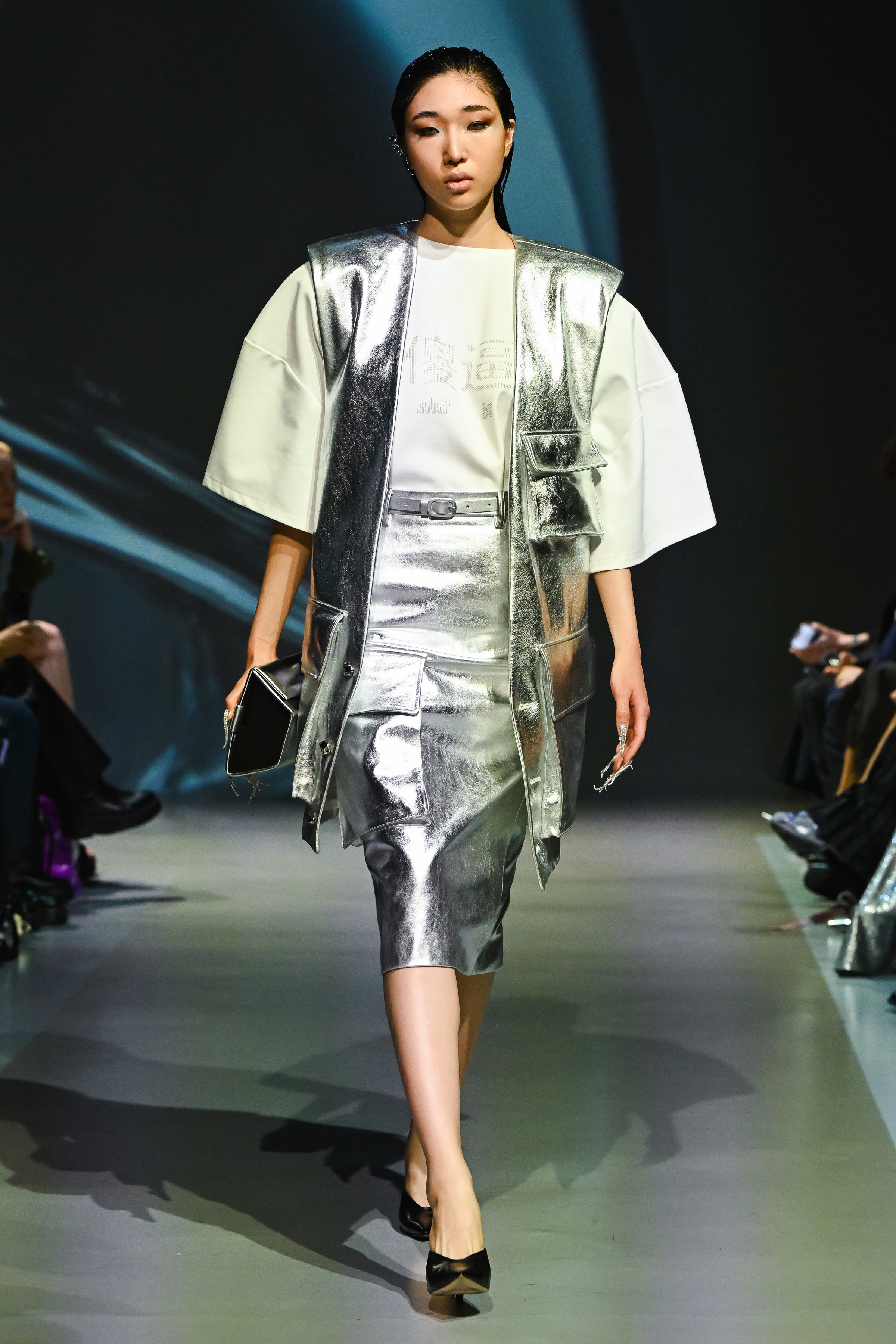 Model walking runway wearing a silver metallic skirt and matching sleeveless vest with sculptural shoulders, holding the SHAO X Dagne Dover Silver Metallic Eyewear Clutch Bag, showcasing luxury designer fashion with a sharp silhouette.