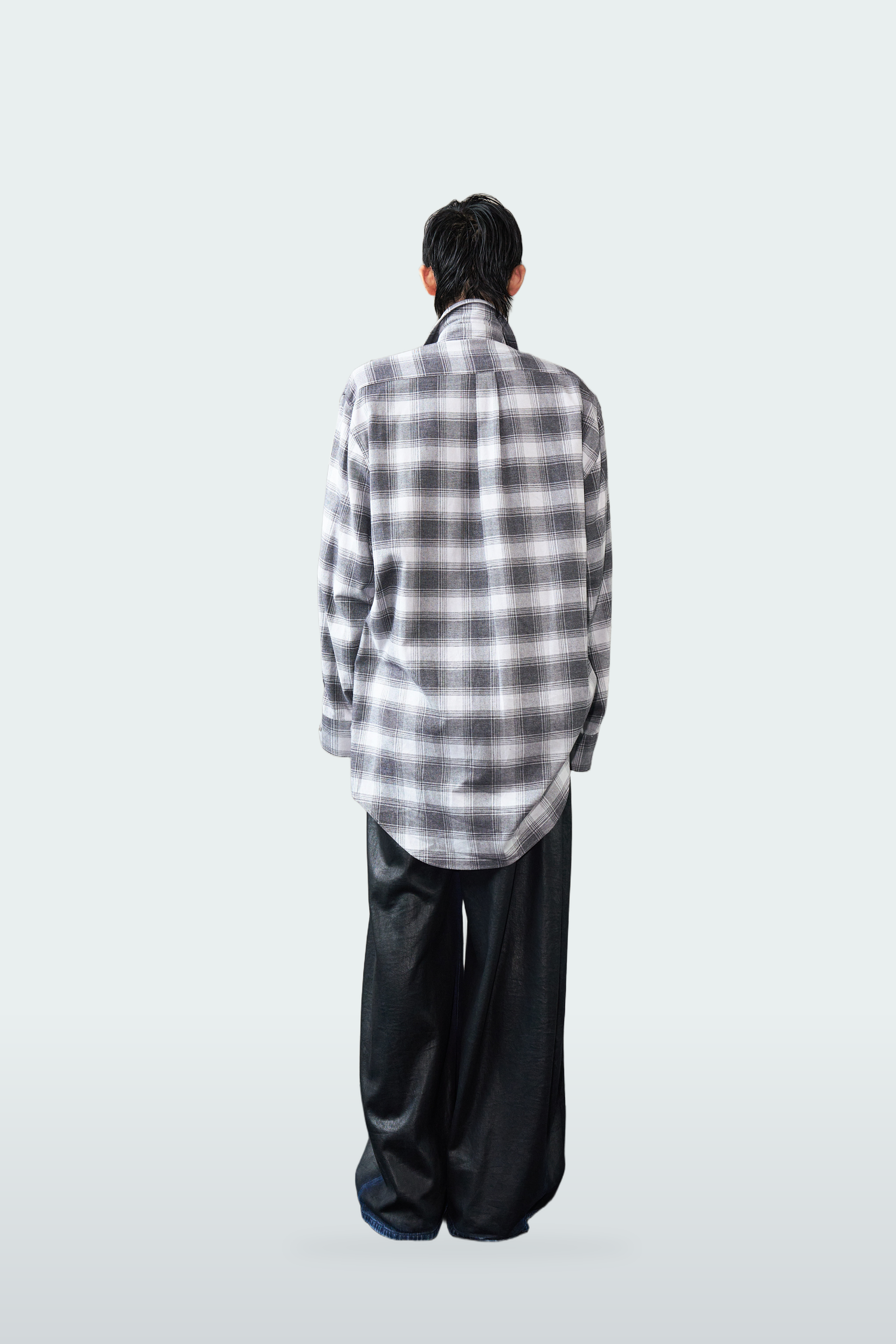 A person viewed from behind wearing an oversized black and white plaid button-up shirt and loose black pants, standing against a plain light gray studio background.