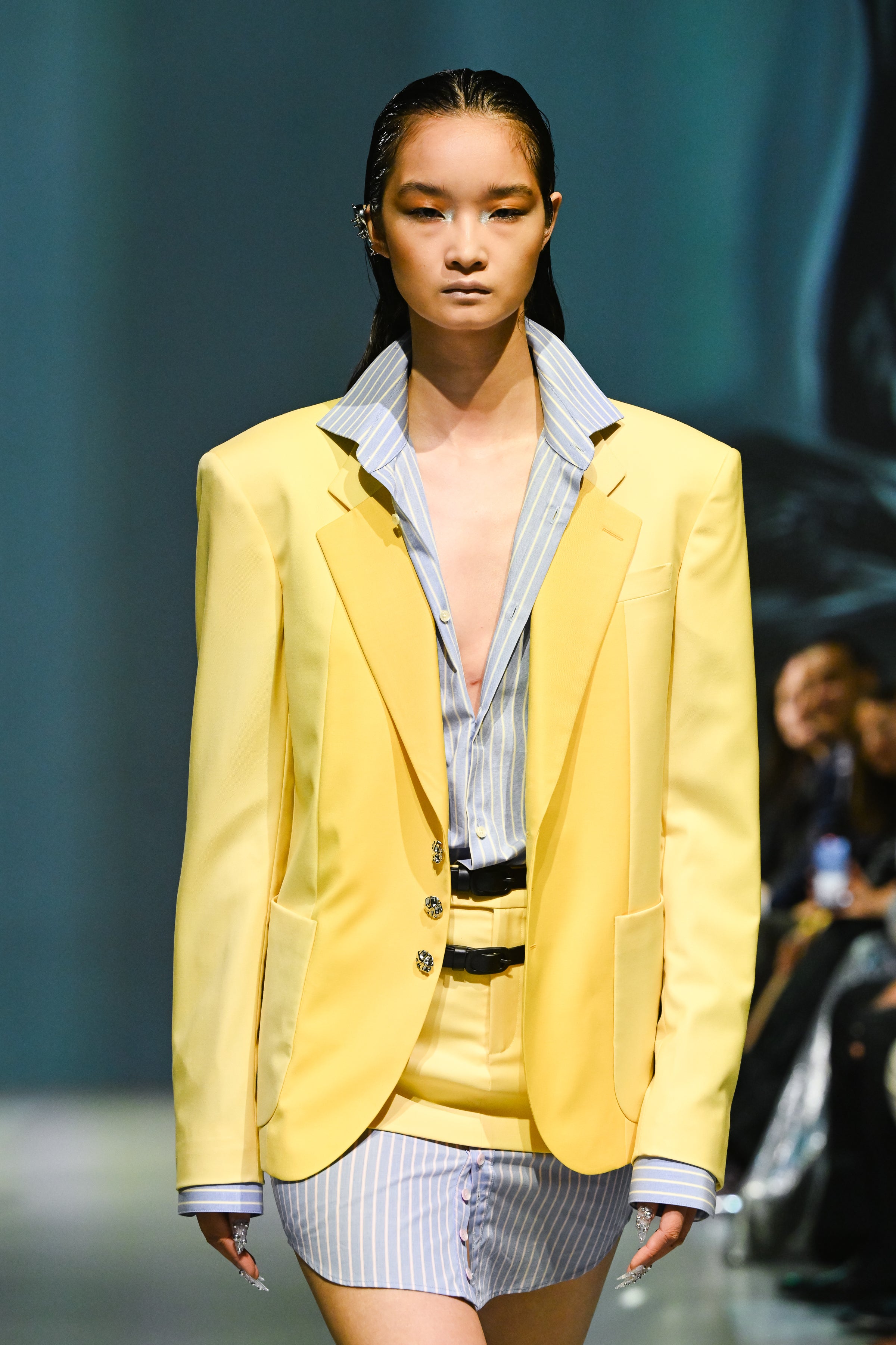 Runway model wearing a structured yellow blazer over a blue and white striped shirt dress with a sculptural silhouette, showcasing avant-garde luxury designer fashion.