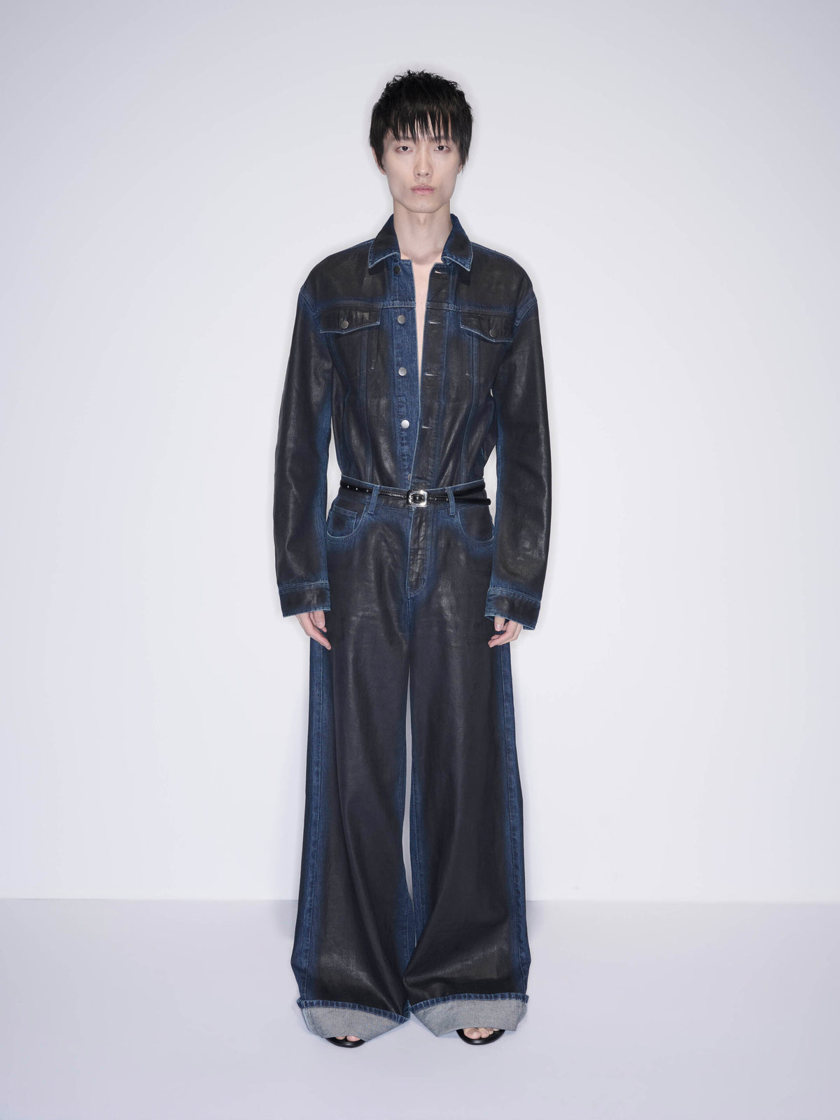 A model stands against a plain light gray studio background wearing a dark denim one-piece jumpsuit with wide legs, a button front, and a matching belt.