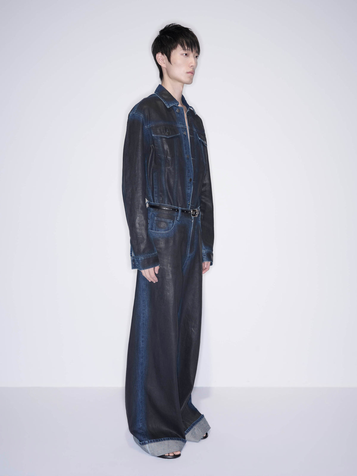 A model stands in profile wearing a dark washed denim one-piece jumpsuit with wide-leg pants, a long-sleeve shirt-style top, and a thin belt, photographed against a plain light gray studio background.