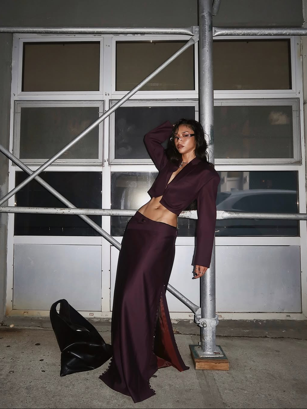 A woman poses outdoors at night wearing a deep burgundy cropped blazer and matching long skirt with a high slit, standing next to metal scaffolding in an urban setting.
